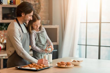 Küçük kız mutfakta babasıyla yemek yerken bardaklara süt dolduruyor, ev yapımı hamur işlerinin tadını çıkarıyor.