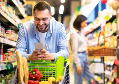 Gülümseyen Erkek Müşteri Süpermarkette Market Alışverişi yaparken Akıllı Telefon Yürüyüşü yapıyor. Seçici Odaklanma. Markette Market Alışveriş Uygulamasını Telefonla Kullanan Adam Yemek Pişiriyor
