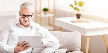 Checking Email. Close up portrait of mature man holding digital tablet sitting on the sofa at home, free space