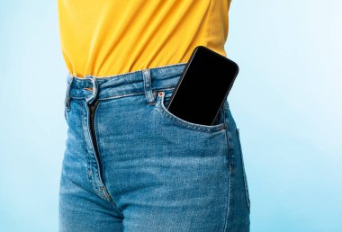 Mavi stüdyo arka planında pantolonunun cebinde boş ekranı olan akıllı telefonu olan tanınmayan bir kadın. Mobil uygulama ya da web sitesi tasarımı için model. İlan şablonunuz için boşluk