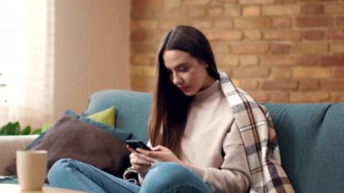 Genç bir kadın, rahat bir oturma odasında, akıllı telefonuna odaklanmış bir kanepede rahatça oturuyor. Yumuşak doğal ışık pencereden süzülerek sıcak bir ortam yaratır..