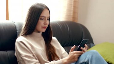 Genç bir kadın, akıllı telefonuna odaklanmış bir kanepede rahatça oturur. Yumuşak doğal ışık pencereden süzülüp yaşam alanında sıcak ve davetkar bir atmosfer yaratıyor..