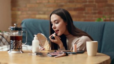 Genç bir kadın, tahta bir masada otururken küçük bir aynanın önünde ruj sürmeye odaklanır. Çeşitli makyaj ürünleri ve kahve makinesi sıcak, davetkar bir odaya yakın otururlar..