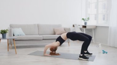 Bir kadın güneşli bir oturma odasında yoga minderinde köprü pozu veriyor. Boşlukta rahat bir kanepe, bitkiler ve büyük pencerelerden gelen doğal ışık var..