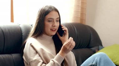 Genç bir kadın rahat bir şekilde kanepede oturuyor, akıllı telefonuyla gevşek bir ifadeyle konuşuyor. Konforlu oturma odasında pencereden süzülen doğal ışık var..