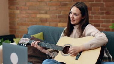 Genç bir kadın dizüstü bilgisayarına bakarken keyifle gitar çalar. Sıcak, davetkar bir oturma odasında, tuğla bir duvar ve yumuşak yastıklarla rahat bir şekilde oturur..