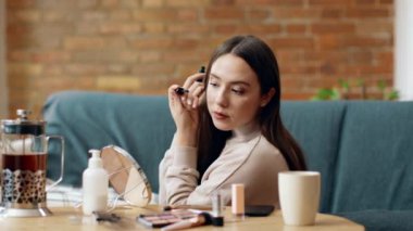 Bir kadın, iyi aydınlatılmış bir odada güzellik ürünleriyle dolu bir masada otururken maskara sürüyor. El kamerasındaki yansımasına odaklanıyor, makyajını geliştiriyor..