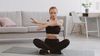 Genç bir kadın yoga minderinin üzerinde bağdaş kurup parlak, modern bir oturma odasında esneme egzersizi yapıyor. Doğal ışık boşluğu suyla doldurur, dingin atmosferi güçlendirir..