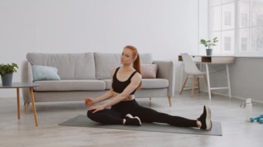 Aktif kıyafet giymiş bir kadın şık bir oturma odasında yoga minderinde esneme egzersizi yapıyor. Parlak alanda bir kanepe, bitkiler ve egzersiz ekipmanları yer alıyor..