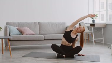 Bir kadın, modern oturma odasındaki paspasın üzerinde yoga yapıyor. Güneş ışığı pencereden süzülüyor, alıştırmasına odaklanırken huzurlu atmosferi arttırıyor..