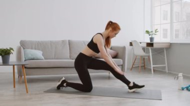 Atletik giyinen bir kadın şık, aydınlık bir yaşam alanında bacak esneme hareketi yapar. Modern dekor bir kanepe, bir masa ve bitkiler içeriyor, egzersiz için sakin bir ortam yaratıyor..