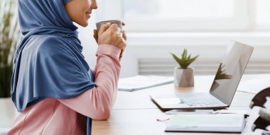 Coffee at work. Relaxed muslim female manager sitting with mug in hands at workplace in office, cropped image, panorama with empty space