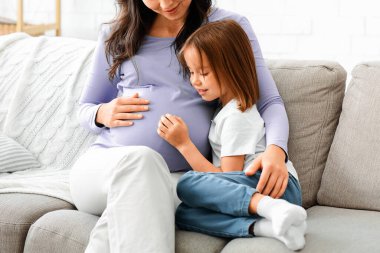 Meraklı küçük kız hamile annesinin karnını gözlemliyor. Evin içi, boş yer.