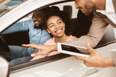 Otomobil galerisinde oturan yeni araba testi yaptıran ve satış temsilcisiyle konuşan mutlu siyah çift. Aile Otomobili Seçimi.