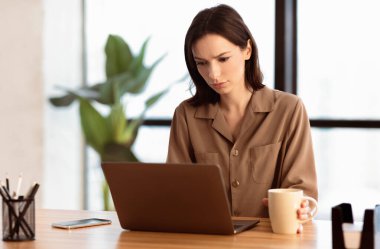 Monoton çalışmaktan yoruldum. Modern ofiste dizüstü bilgisayarla çalışan, üzgün hisseden, elinde kahve fincanı tutan zavallı bir kız.