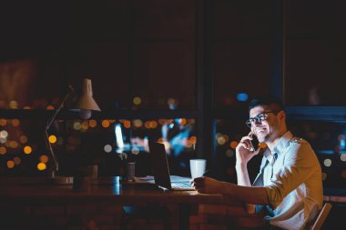 Başarılı bir iş adamı telefonda konuşuyor. Geceleri modern ofiste pencerenin yanında çalışıyor. Boşluğu Kopyala