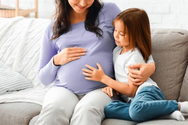 Mutlu hamile anne ve anaokulu kızı evdeki kanepede oturuyor, sohbet ediyor, boşluğu kopyalıyor.