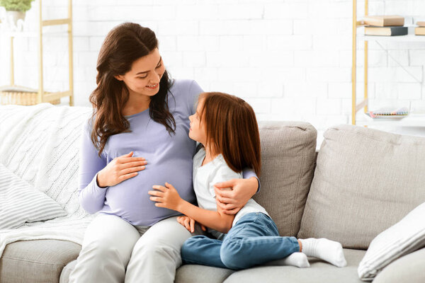 Pregnant woman and little daughter sitting on sofa at living room and having conversation, free space