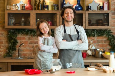 Ev Fırıncıları. Mutlu genç baba ve küçük kızın portresi, mutfak önlüklerinde poz vermeleri, ev yapımı hamur işlerinin tadını çıkarmaları, birlikte hamur işlerini hazırlamaları, kollarını kavuşturup kameraya gülümsemeleri,