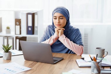İş Planlama Konsepti. Düşünceli Müslüman iş kadını iş yerinde oturmuş, düşünceli yüz ifadesiyle dizüstü bilgisayar ekranına bakıyor.