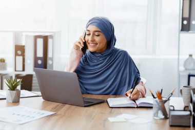 İş hayatı tarzı. Ofisteki meşgul Orta Doğulu iş kadınının portresi, cep telefonuyla konuşmak, dizüstü bilgisayar kullanmak ve not almak.