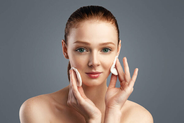 Рыжеволосая девушка чистит кожу с хлопчатобумажными прокладками, глядя на камеру, позирующую, заботясь о лице над белым фоном студии. Кожа лица и косметика, концепция чистой кожи. Портрет красоты