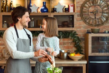 Neşeli genç baba ve küçük kız öğle yemeği için sebze salatası pişiriyor, bowlinge baharat ekliyor, ev yemeğinin tadını çıkarıyor, evde eğleniyor, önlük giyiyor ve gülümsüyor ve boş alan yaratıyor.