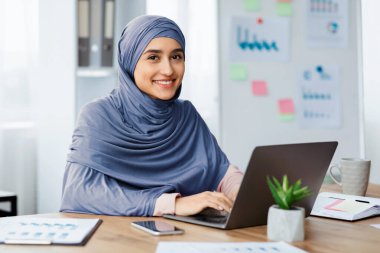 Modern ofiste masa başında oturan, dizüstü bilgisayar kullanan ve kameraya gülümseyen tesettürlü güzel Arap kadın. Seçici odaklı portre