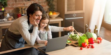 Neşeli bekar baba ve küçük kızı dizüstü bilgisayarlarını kullanıyorlar yemek malzemelerini kontrol ediyorlar, mutfakta birlikte yemek pişiriyorlar.
