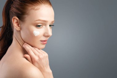 Yüzüne kalp şeklinde krem sürmüş, beyaz stüdyo geçmişine bakan şehvetli kızıl saçlı genç bayan. Gençlik Konsepti için Yüz Cilt Kozmetiği