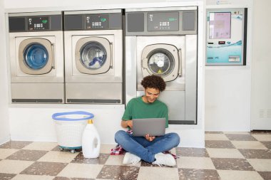 Genç bir adam çamaşırhanenin zemininde oturuyor, dizüstü bilgisayarına odaklanmış, arka planda çamaşır makineleri var. Kıyafet beklerken birden fazla iş yaparken rahat ve üretken görünüyor..