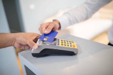 Müşteri, perakende kasasındaki ödeme terminalinden kredi kartıyla ulaşılmaz bir ödeme yapıyor. Kasiyer, parlayan bir mağaza ortamında işleme yardımcı olur..