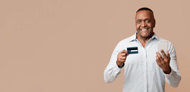 Çevrimiçi bankacılık kavramı. Neşeli Afro-Amerikalı adam kart dengesini mobil uygulama ve turuncu stüdyo geçmişi ile kontrol ediyor.