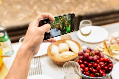 Bir el, açık havada aile toplantısı sırasında grup selfie 'sini çeken bir akıllı telefonu tutuyor. Masada meyve ve ekmek gibi lezzetli yiyecekler sergileniyor, neşeli atmosferi güçlendiriyor..