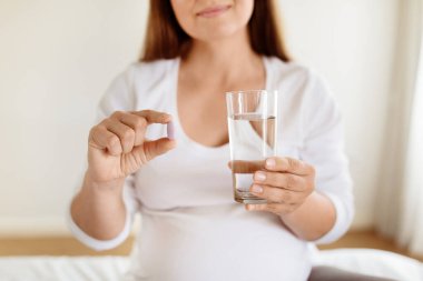 Hamile bir kadın, bir elinde bir bardak su, diğer elinde de ilave almaya hazırlanan aydınlık bir yerde oturur. Sakin ve sağlığına odaklanmış görünüyor..