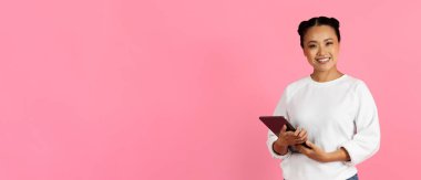 Neşeli genç bir kadın kendinden emin bir şekilde elinde tabletle ayakta duruyor ve sıcak bir şekilde gülümsüyor. Canlı pembe bir zemin üzerinde gelişigüzel giyinmiş. Olumlu ve yaratıcılık, kopyalama alanı öneriyor.