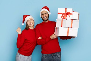 Kırmızı süveter ve Noel Baba şapkası giymiş mutlu bir çift neşeli mavi bir arka planda kurdelelerle paketlenmiş hediye kutularını birlikte tutuyorlar..