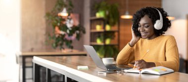 Kablosuz kulaklıklı mutlu siyah kız çevrimiçi çalışıyor, dizüstü bilgisayar kullanıyor ve notlar alıyor, kafenin içini ve kopyalama alanını kullanıyor.