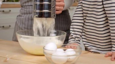 Bir çocuk, sıcak bir mutfakta yemek tarifi hazırlarken bir ebeveynin unu karıştırma kabına koymasını izler..