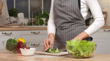 Bir kadın sebze ve sebzelerle dolu şık bir mutfakta salata ve salatalık doğrayarak taze bir salata hazırlıyor..