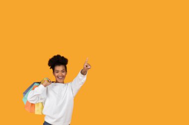 Beyaz kazak giyen neşeli bir kadın heyecanlı bir şekilde birçok renkli alışveriş torbası tutuyor. Canlı turuncu arka plan alışverişte onun neşeli yüz ifadesine ekleniyor..