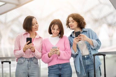 Üç arkadaş birbirine yakın dururlar, akıllı telefonlarına dalmış bir şekilde, kapalı bir alanda aydınlık bir anı paylaşırlar, muhtemelen bir havaalanında veya modern bir binada..
