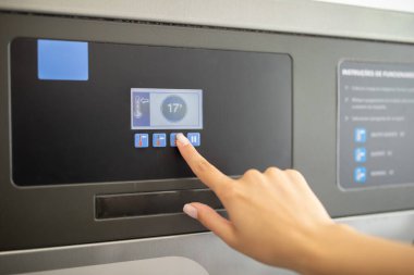 Bir kişi dokunmatik ekran kontrol panelindeki düğmelere basıyor, muhtemelen bir tesisteki ayarları ayarlıyor. Arayüz sıcaklık ve ek seçenekleri açıkça gösterir.