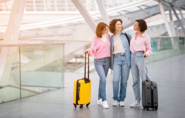 Üç arkadaş, bavullarını parlak ve ferah bir havaalanı terminalinde tutarken birlikte gülüyor ve gülümsüyor ve seyahat için birliklerini ve heyecanlarını sergiliyorlar..