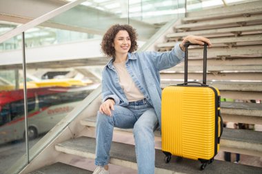 Kıvırcık saçlı neşeli bir kadın işlek bir terminalin merdivenlerinde parlak sarı bavulunun yanında dinleniyor. Seyahat koşuşturması ve telaşının ortasında bir anlık dinlenmenin tadını çıkarıyor..