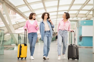 Üç arkadaş birlikte bir ulaşım terminalinde yürüyorlar, her biri yanında bir bavulla kahkaha ve gülümseme paylaşıyorlar. Güneş ışığı üzerlerindeki cam tavandan süzülüyor..