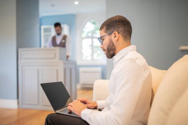 A man wearing glasses and a white shirt sits on a couch in a hotel lobby. He is working on a laptop. A person standing behind a desk in the background is out of focus. clipart