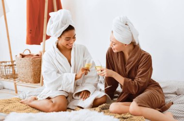 Bornozlu iki neşeli kız arkadaş evde spa yapıyorlar, yatakta oturuyorlar, şampanya içip sohbet ediyorlar.