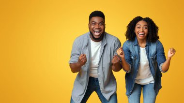 Afro-Amerikan çifti heyecanlı bir şekilde bağırıyor ve sıkılmış yumruklarla tezahürat yapıyor, sarı stüdyonun arka planında duruyor.
