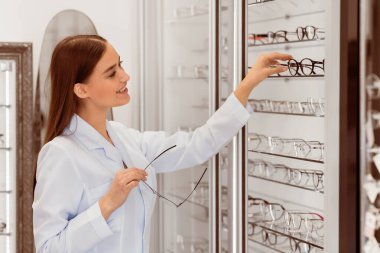 Kadın oftalmolog. Gülümseyen satıcı, optik dükkandaki vitrinde çeşitli göz gözlükleri gösteriyor.
